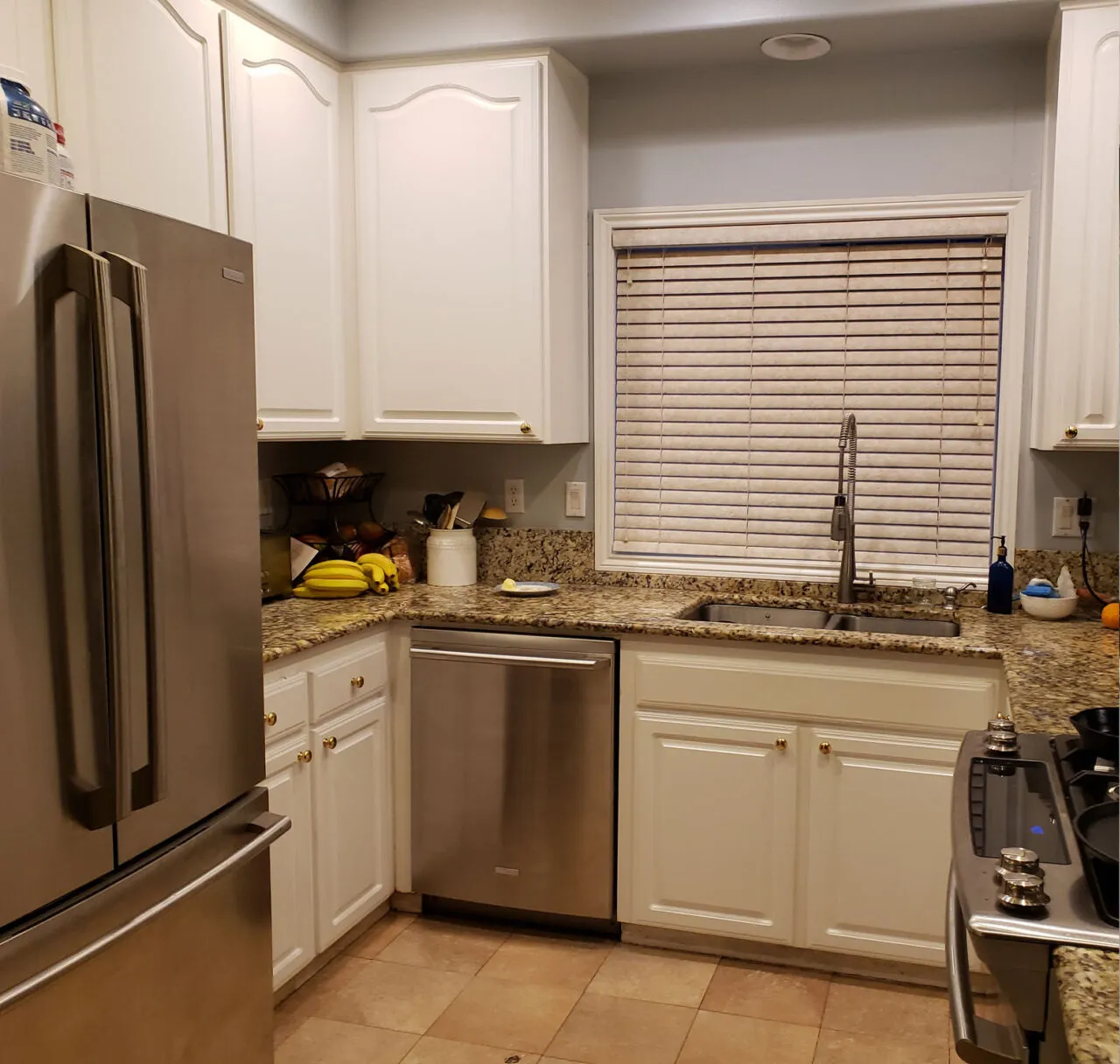 The Davis kitchen before the remodel — cathedral-arch white cabinets, granite counters, mini blinds over the sink and a tile floor