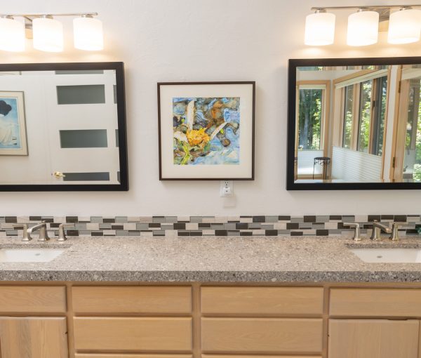Bathroom vanity with warm wood cabinets, long countertop, two mirrors, wall lights, framed artwork, and patterned tile backsplash.