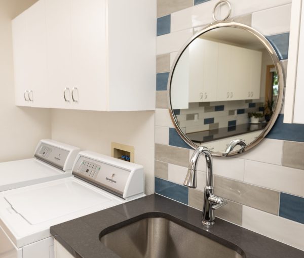 Close-up of laundry room sink with round mirror, white cabinetry, dark quartz countertop, and blue, gray, and white tile backsplash.