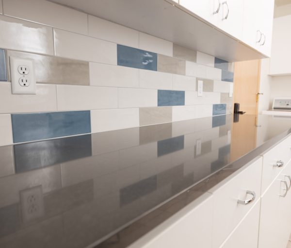 Long quartz countertop in a laundry room with white cabinetry, blue and gray tile backsplash, and washer or dryer visible in the background.