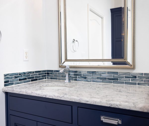 Close-up of bathroom vanity with dark blue cabinets, stone countertop, blue tile backsplash, sink, faucet, and framed mirror