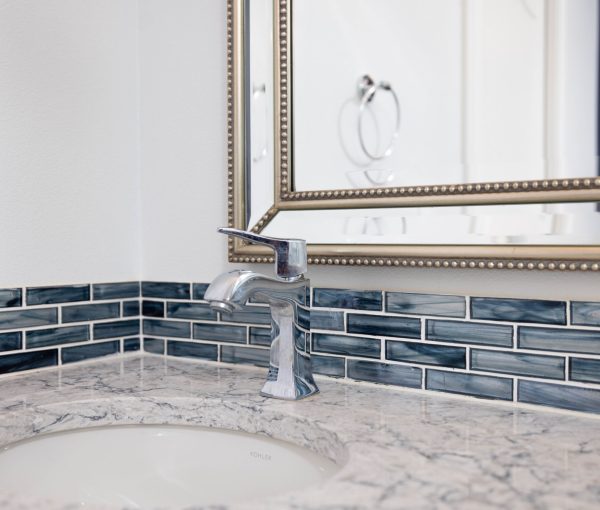 Close-up of bathroom sink with chrome faucet, marble-look countertop, blue tile backsplash, and framed mirror.