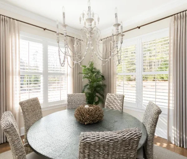 Dining room with round table, woven dining chairs, chandelier, large windows, and plant centerpiece.