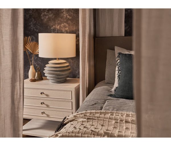 Bedroom detail with bedside table, table lamp, layered bedding, dark drapery, and textured pillows.