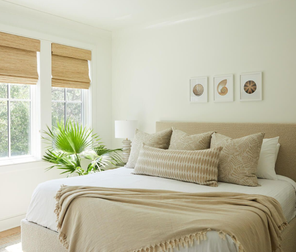 Neutral bedroom with light bedding, layered pillows, woven window shades, potted plant, and simple wall art.