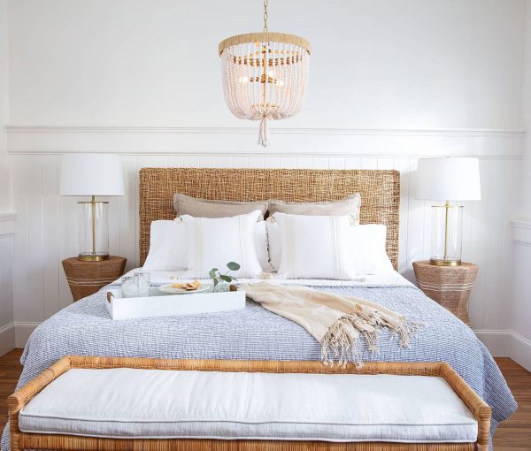 Bedroom with woven headboard, white bedding, wood nightstands, table lamps, chandelier, and layered throw blanket.