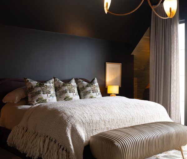 Bedroom with dark accent wall, upholstered bed, layered bedding, bedside lamp, and drapery.